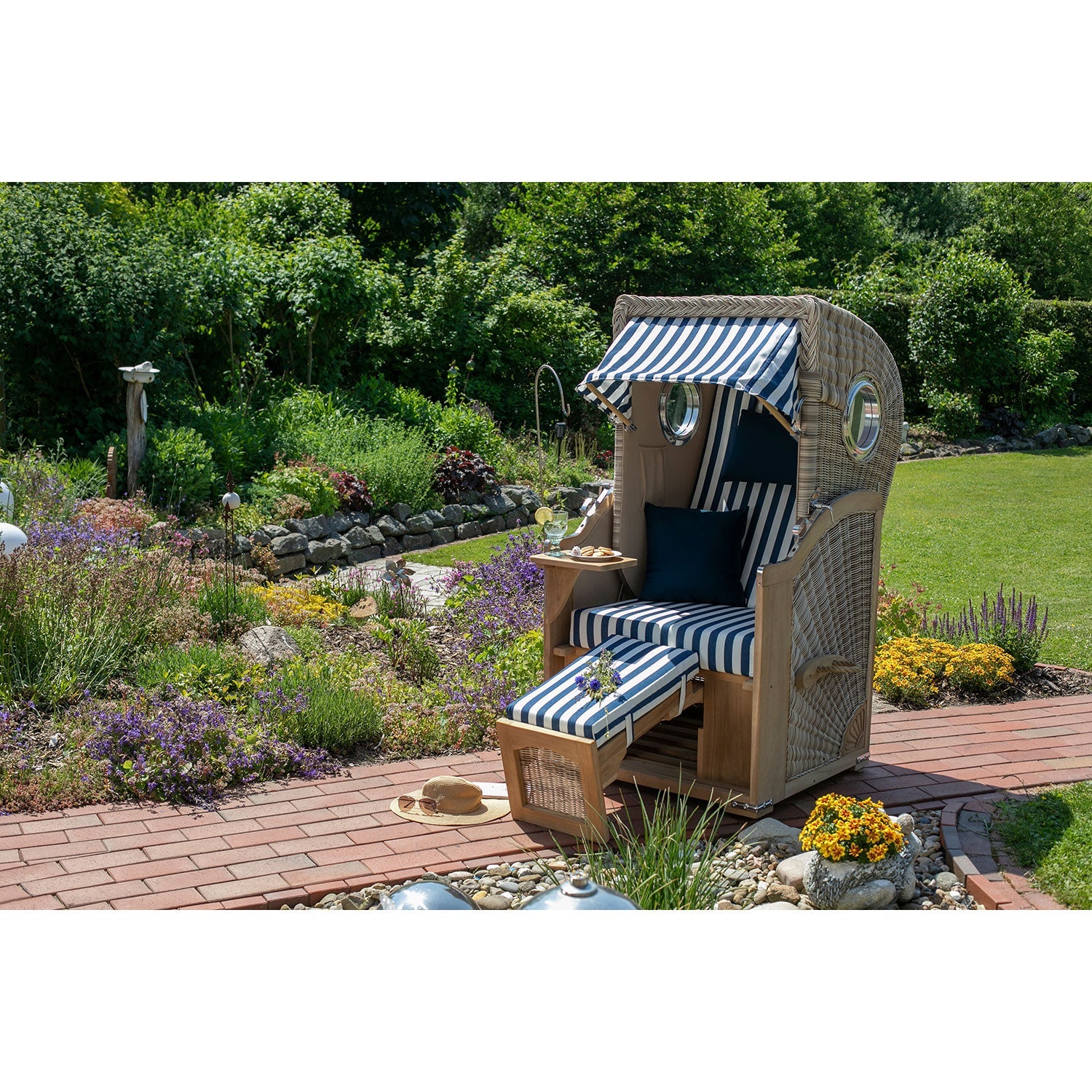 Ein Strandkorb klein – Düne Design 518 mit blau-weiß gestreiften Kissen, einer integrierten Fußstütze und einem Baldachin steht auf einem gepflasterten Weg in einem üppigen Garten. In der Nähe befinden sich Pflanzen, ein Sonnenhut und eine hölzerne Vogelskulptur. Im Hintergrund erstreckt sich ein grüner Rasen.