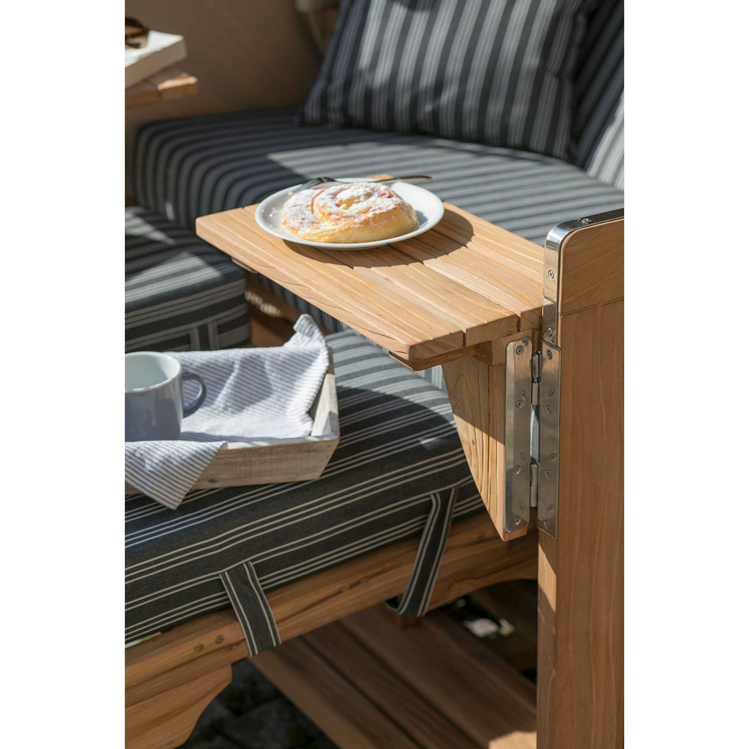 Nahaufnahme eines ausklappbaren Holztisches, der an den gestreiften Außensitzen des Strandkorbs DÜNE, grau mit Nadelstreifen, befestigt ist. Ein Teller mit Gebäck und einer Gabel steht auf dem Tisch, während darunter ein Tablett mit einer Tasse und einem Tischtuch auf den Kissen ruht und diesen gemütlichen Rückzugsort am Strand unterstreicht.