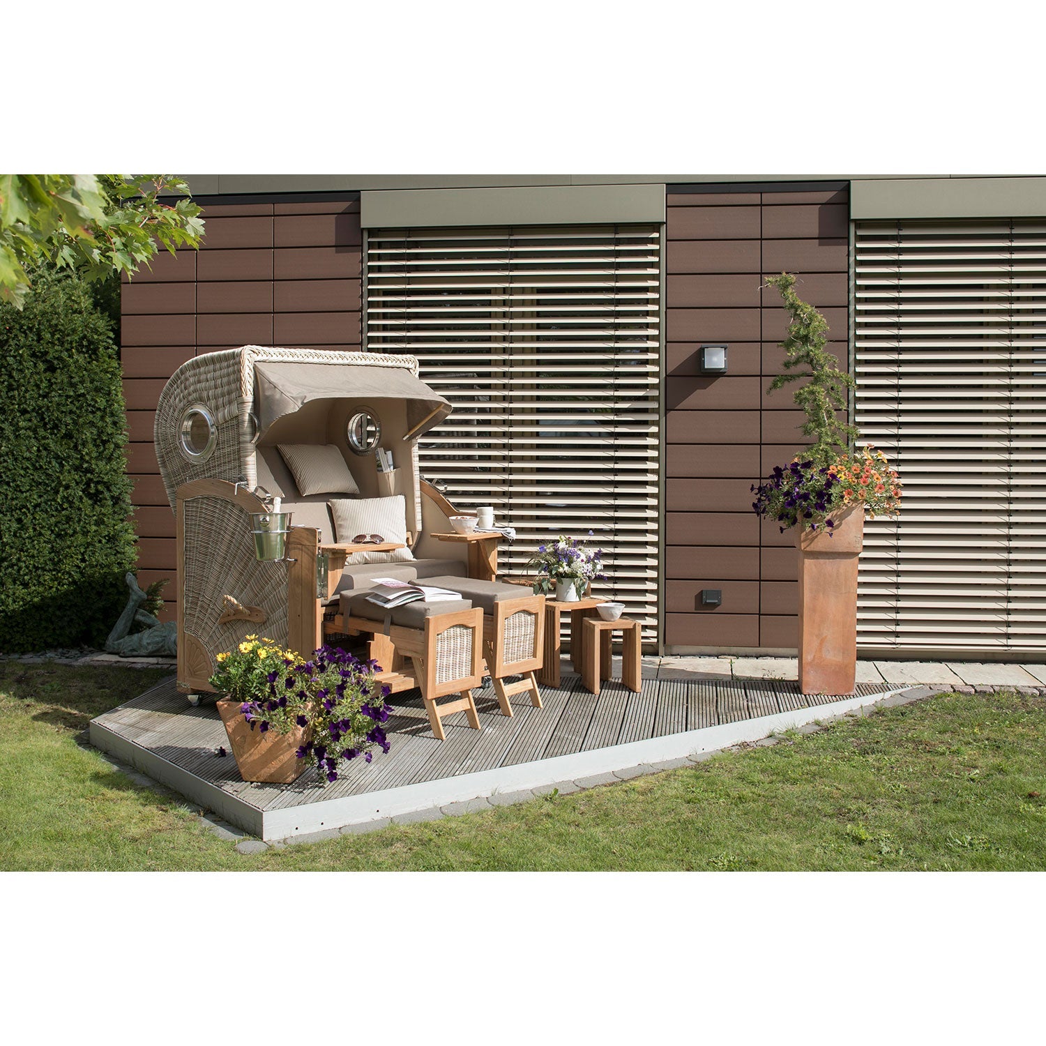 Im Hinterhof steht der gemütliche Strandkorb DÜNE in Taupe-Creme mit Kissen auf einer Holzterrasse. Er besteht aus PE-Geflecht und ist mit Fußstützen und zwei kleinen Tischen ausgestattet. Umgeben von Topfblumen steht er. Dahinter befinden sich Fenster mit großen Holzläden, üppiges Gras und eine angrenzende Hecke.