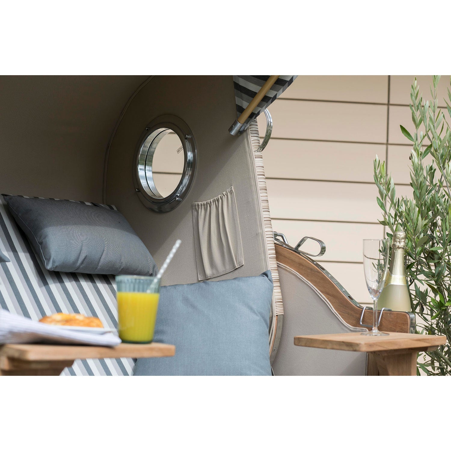 Eine Sitzecke im Freien besticht durch Teakholz-Akzente und gestreifte Kissen. Der Strandkorb DÜNE in grau gestreift verleiht dem Raum einen maritimen Charme. Auf dem Tisch daneben steht ein Tablett mit Champagner und einem Glas Orangensaft mit Strohhalm, ergänzt durch Grünpflanzen im Hintergrund.
