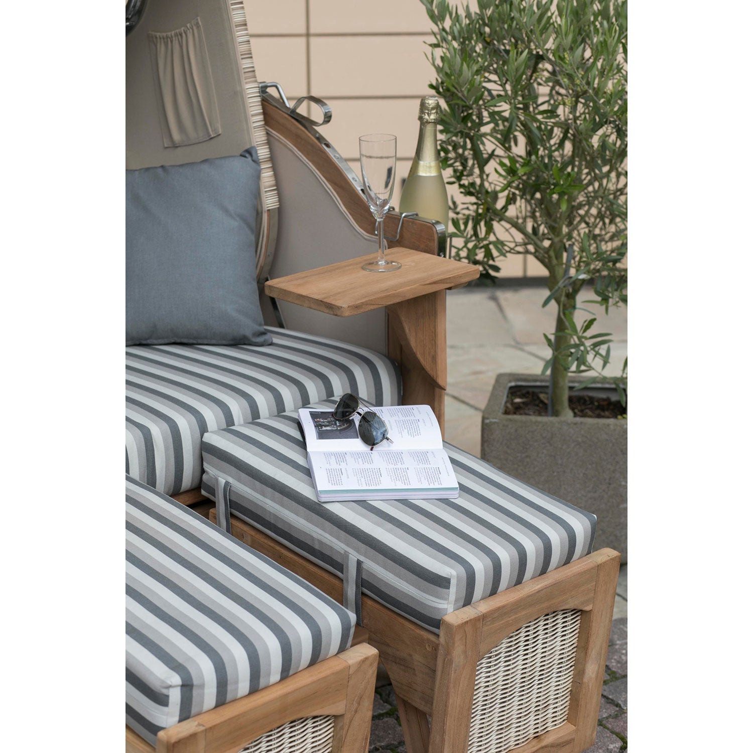 Ein gemütliches Outdoor-Setting besteht aus einem gestreiften Kissen auf einem Strandkorb DÜNE, grau gestreift, gepaart mit einem Beistelltisch mit Champagner. Daneben stehen ein Buch, eine Sonnenbrille und eine Topfpflanze vor einer strukturierten Wand für eine ruhige Szene.