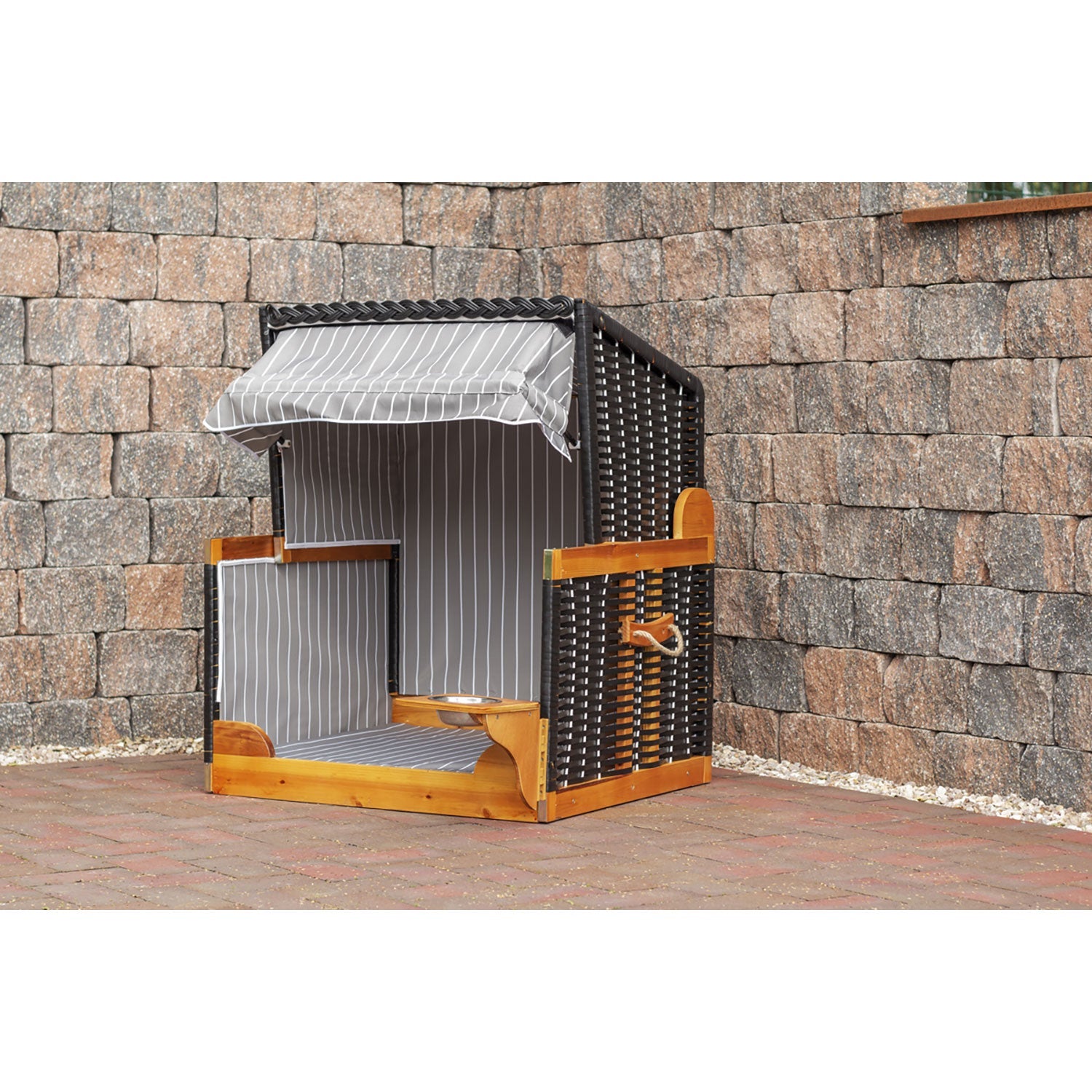 Ein Strandkorb für Hunde, anthrazit, verfügt über einen Korbrahmen aus hochwertigem Wachholderholz und wetterbeständigem PE-Geflecht. Er steht auf einer gemauerten Terrasse vor einer mit kleinen weißen Kieselsteinen gesäumten Steinmauer.