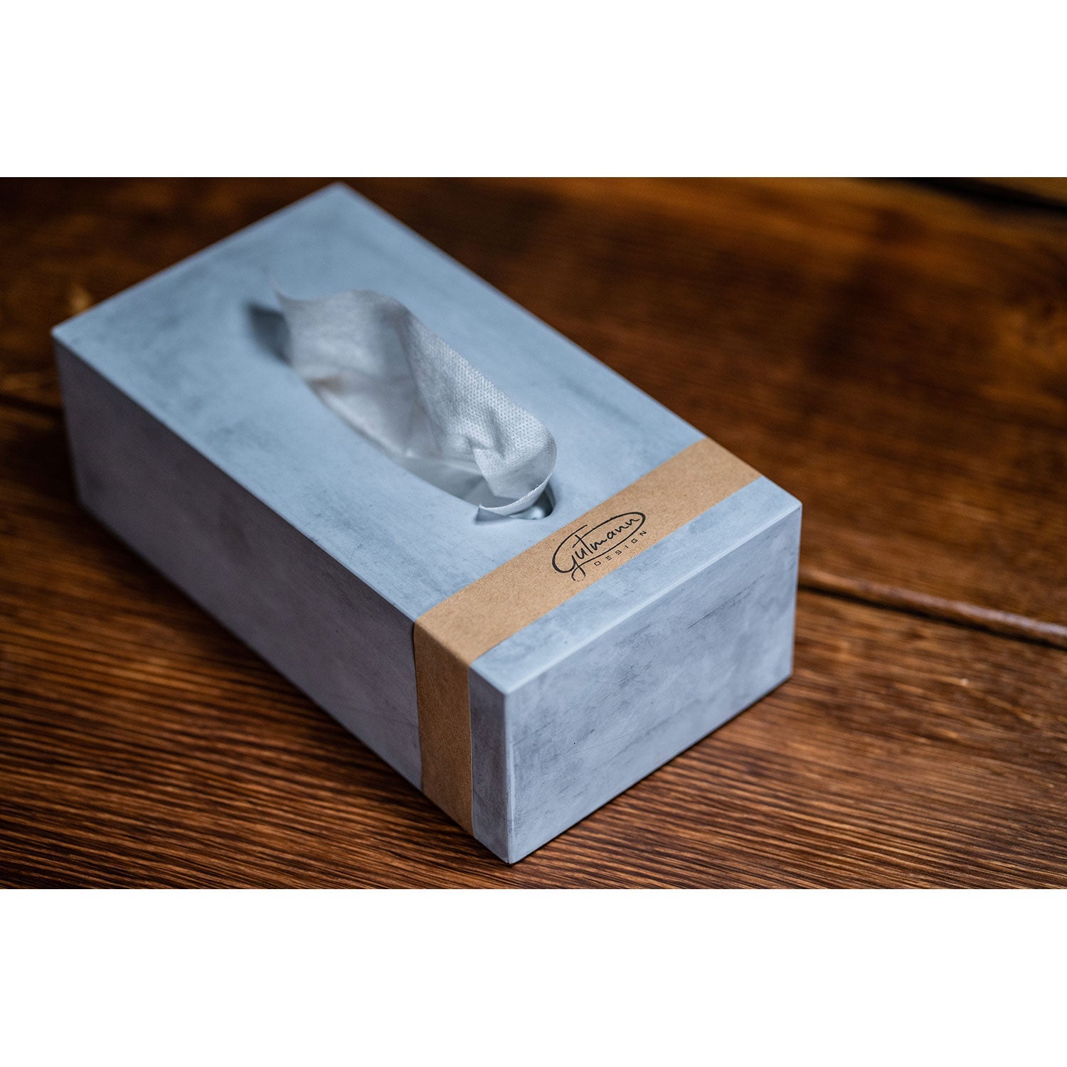 Eine Taschentücherbox aus Beton mit grauer Oberfläche und einem hervorstehenden Taschentuch steht auf einem Holztisch. Das braune Band mit Text unterstreicht die moderne Ästhetik und betont den Kontrast zwischen der glatten Oberfläche und dem strukturierten Holz.