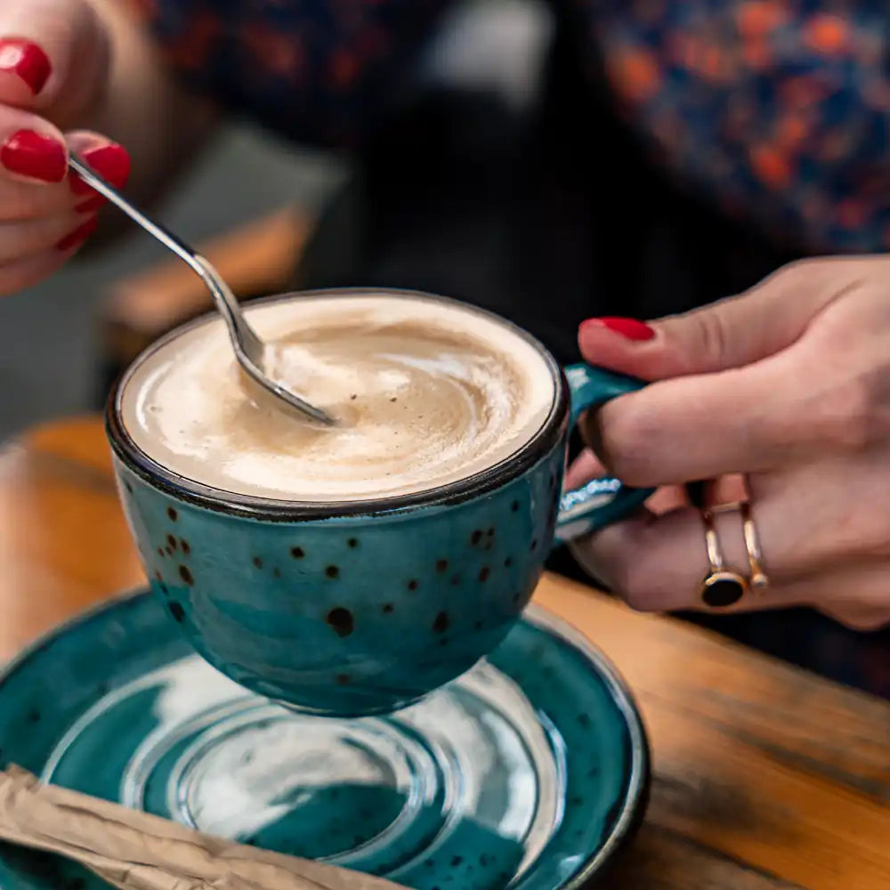 Person, die eine Tasse Kaffee mit einem Löffel umrührt und einen blauen Becher mit roten Nägeln hält.
