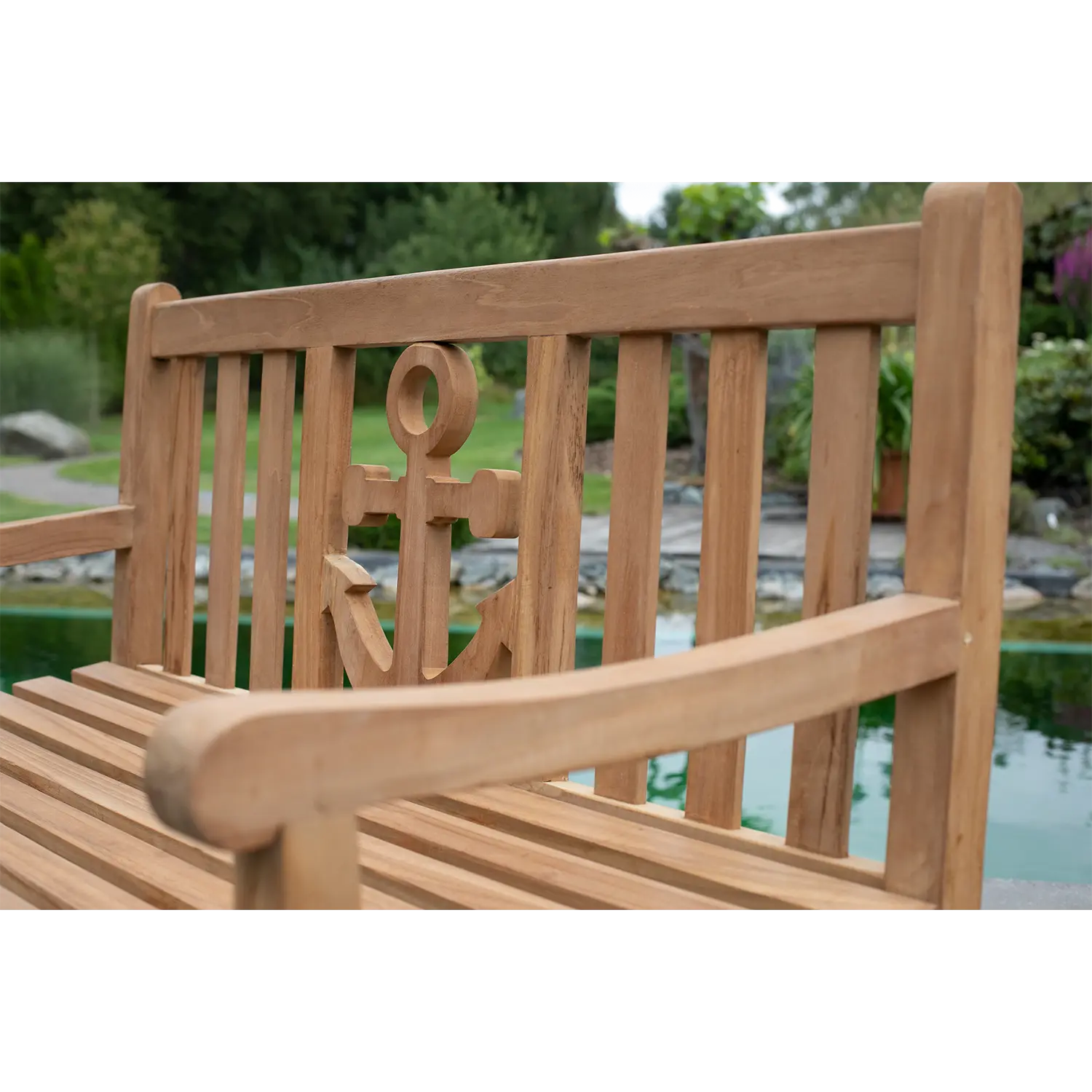 Eine Nahaufnahme der Rückenlehne der Teak-Gartenbank Anker unterstreicht ihr unverwechselbares Anker-Design. Im Hintergrund ist ein Teich zu sehen, umgeben von üppigem Grün und Felsen, der eine ruhige Outdoor-Atmosphäre schafft.