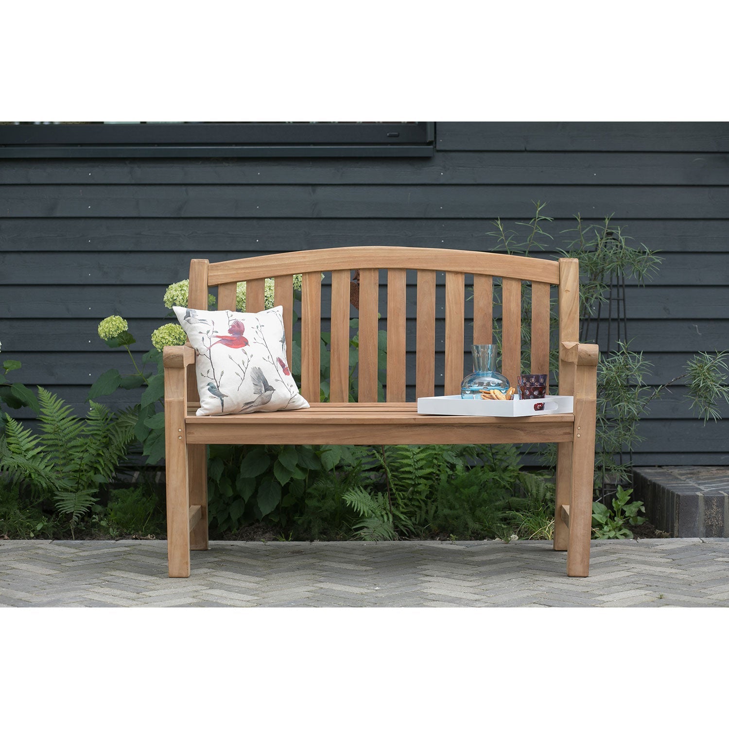 Vor der dunkel getäfelten Wand steht die Gartenbank aus Holz 2-Sitzer mit Pflanzen dekoriert. Auf dem Bankkissen mit rotem Vogelmotiv befindet sich ein Tablett mit Glas und Schälchen – ein stilvolles Outdoor-Highlight.