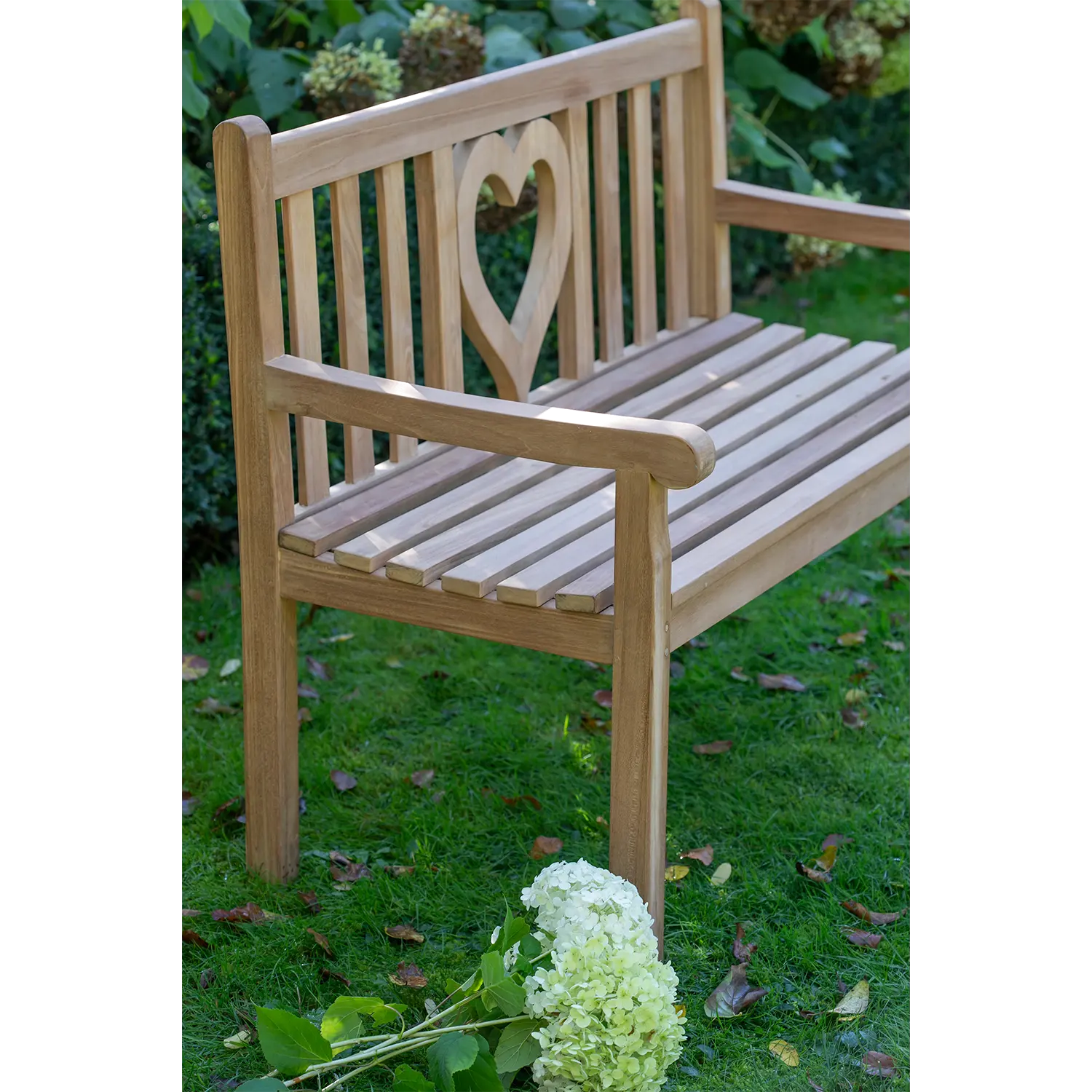 Die Teak-Gartenbank Herz, eine Holzbank mit herzförmigem Ausschnitt in der Rückenlehne aus Teakholz, steht auf Rasen. Daneben zieren weiße Hortensien das Vorderbein dieser charmanten Outdoor-Sitzgelegenheit, umgeben von üppigem Grün im Hintergrund.