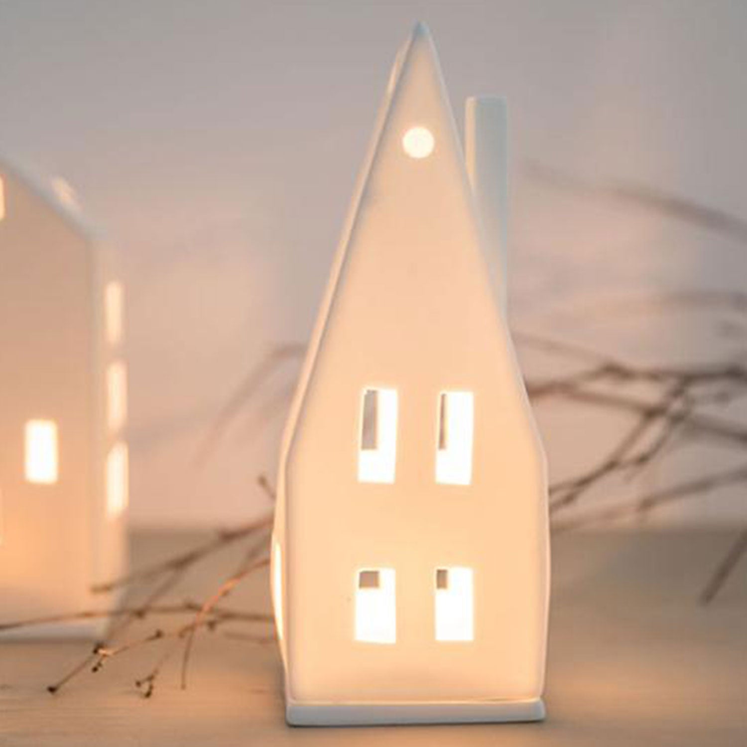 Das Kleine Lichthaus „Spitzdach“ ist ein kleines, weißes Porzellanhaus mit hohem Spitzdach und leuchtenden Fenstern. Zarte Zweige im Hintergrund sorgen für eine warme und gemütliche Weihnachtsstimmung.