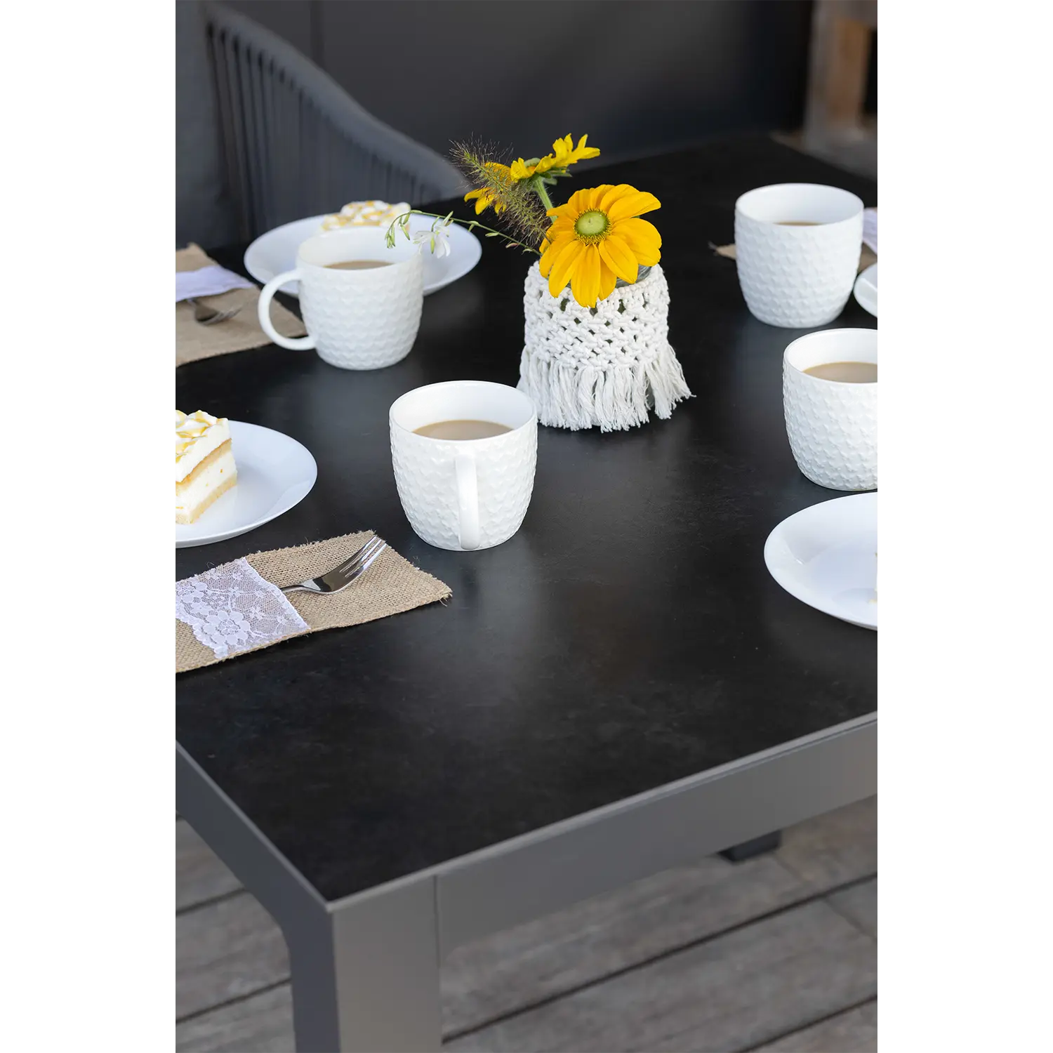 Auf einem schwarzen Tisch Tarifa stehen weiße Keramiktassen mit Kaffee, weiße Teller mit Kuchenstücken, eine Vase mit gelben Blumen in der Mitte und Gedecke mit Spitze auf Juteservietten.