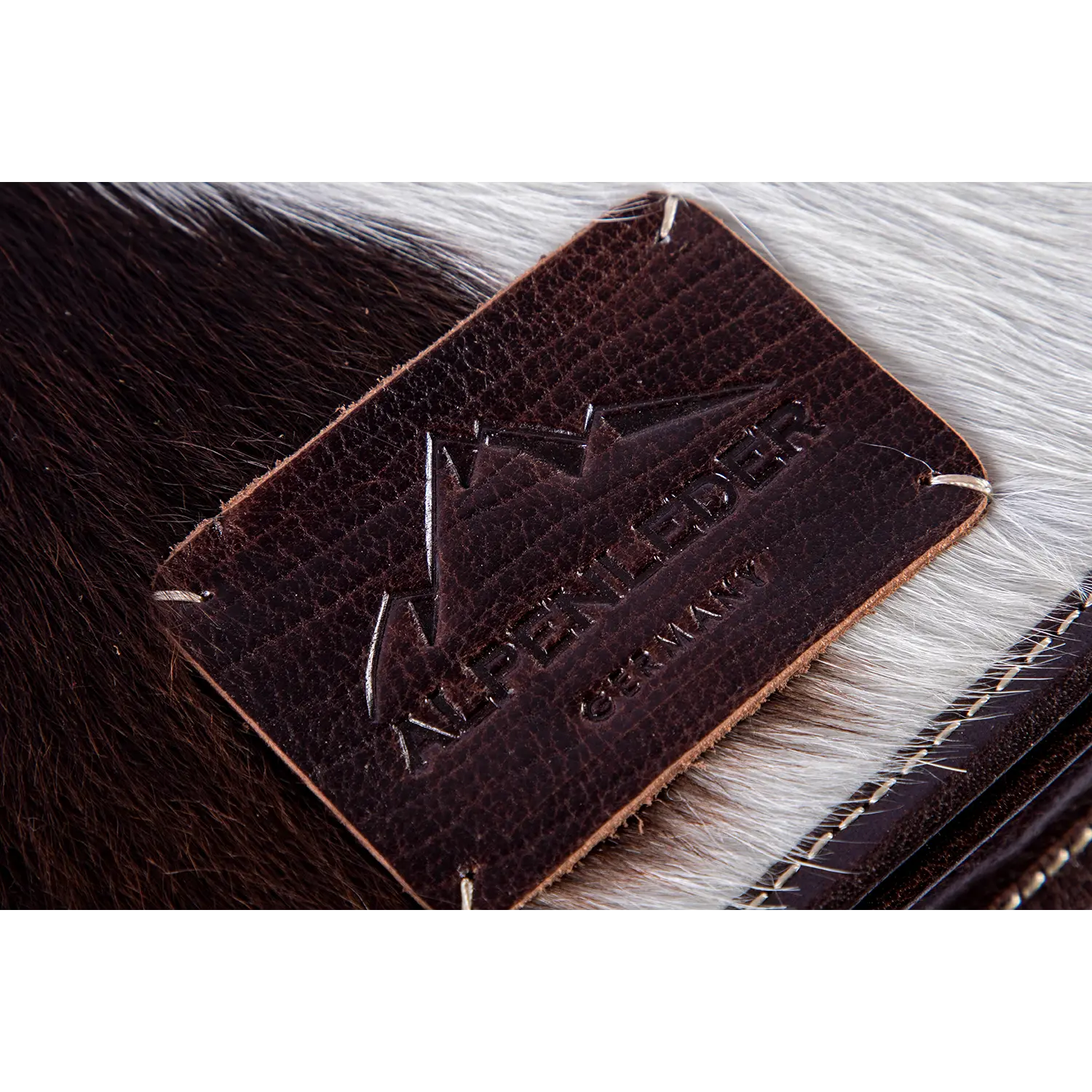 Die Umhängetasche VRONI mit Haaren zeigt die Nahaufnahme eines strukturierten Lederaufnähers mit geprägtem Bergdesign und „Alpenleder“ über „Germany“, sorgfältig auf eine Oberfläche mit Kuhfell genäht, wobei dunkelbraune und weiße Fellstrukturen mit natürlicher Felle verflochten sind.