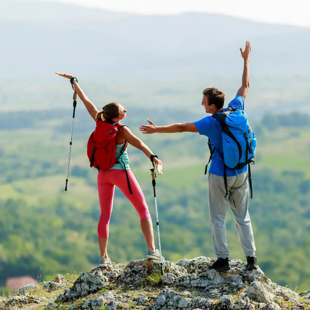 Zwei Wanderer mit Rucksäcken feiern auf einer felsigen Bergkuppe mit erhobenen Armen.