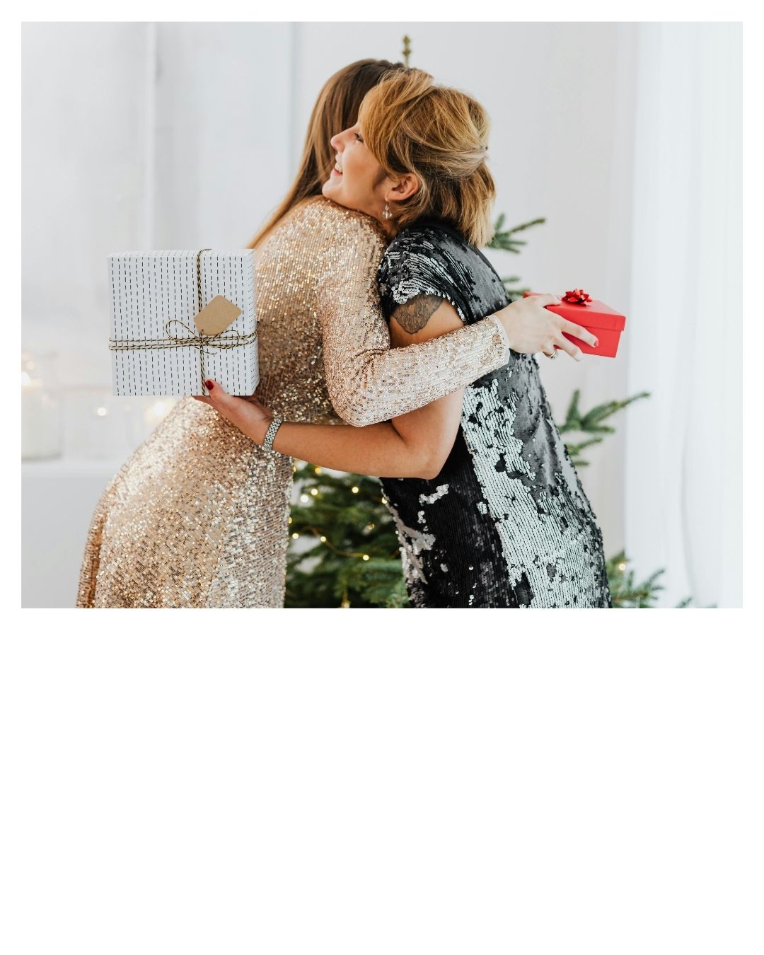 Zwei Frauen in glitzernden Kleidern umarmen sich in der Nähe eines Weihnachtsbaums und halten Geschenke.