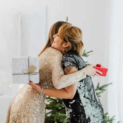 Zwei Frauen in glitzernden Kleidern umarmen sich, während sie eingepackte Weihnachtsgeschenke austauschen.