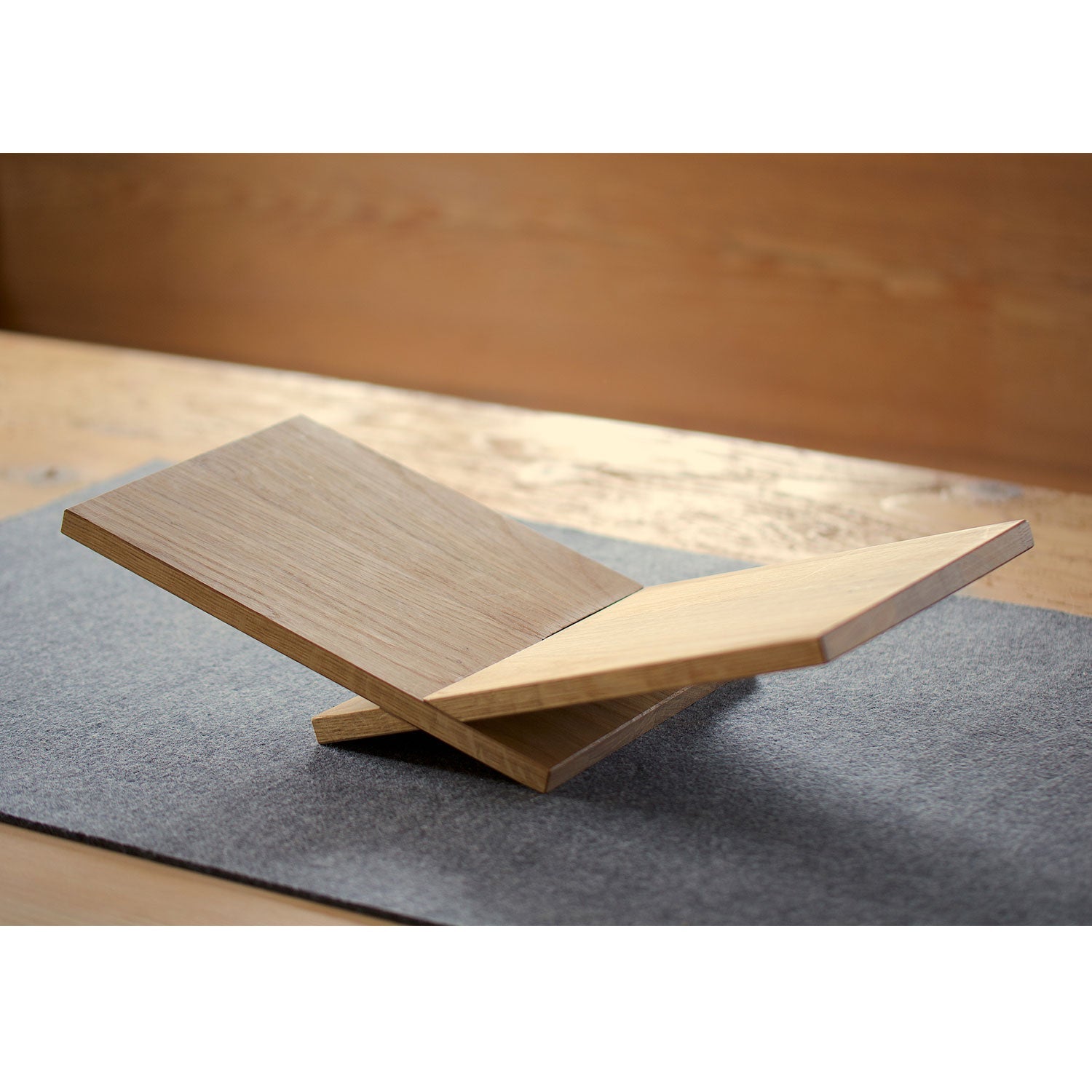 Der hölzerne Bücherständer „Weinliege“ mit seinem schlichten, eckigen Design und den sich kreuzenden Paneelen, die ein X bilden, ruht auf einer grauen Matte auf einer Holzoberfläche. Eine sanft fokussierte Holzwand dient als Hintergrund.