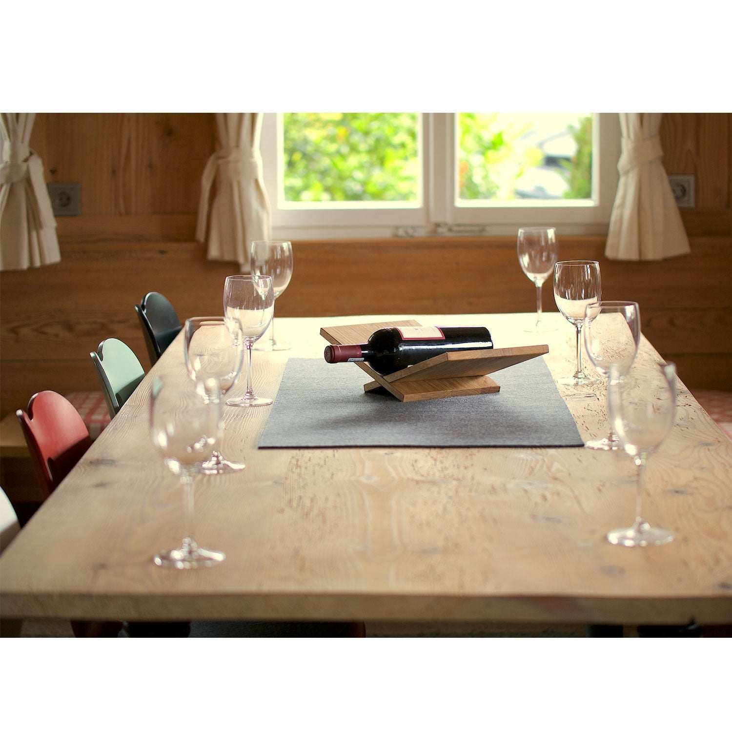 Ein hölzernes Esstischset mit acht leeren Weingläsern und einer Flasche Rotwein in einer eleganten Weinliege. Die hellen Holzwände, der Blick durch das cremefarbene Fenster und die charmante Obstschale schaffen eine elegante Ruhe.