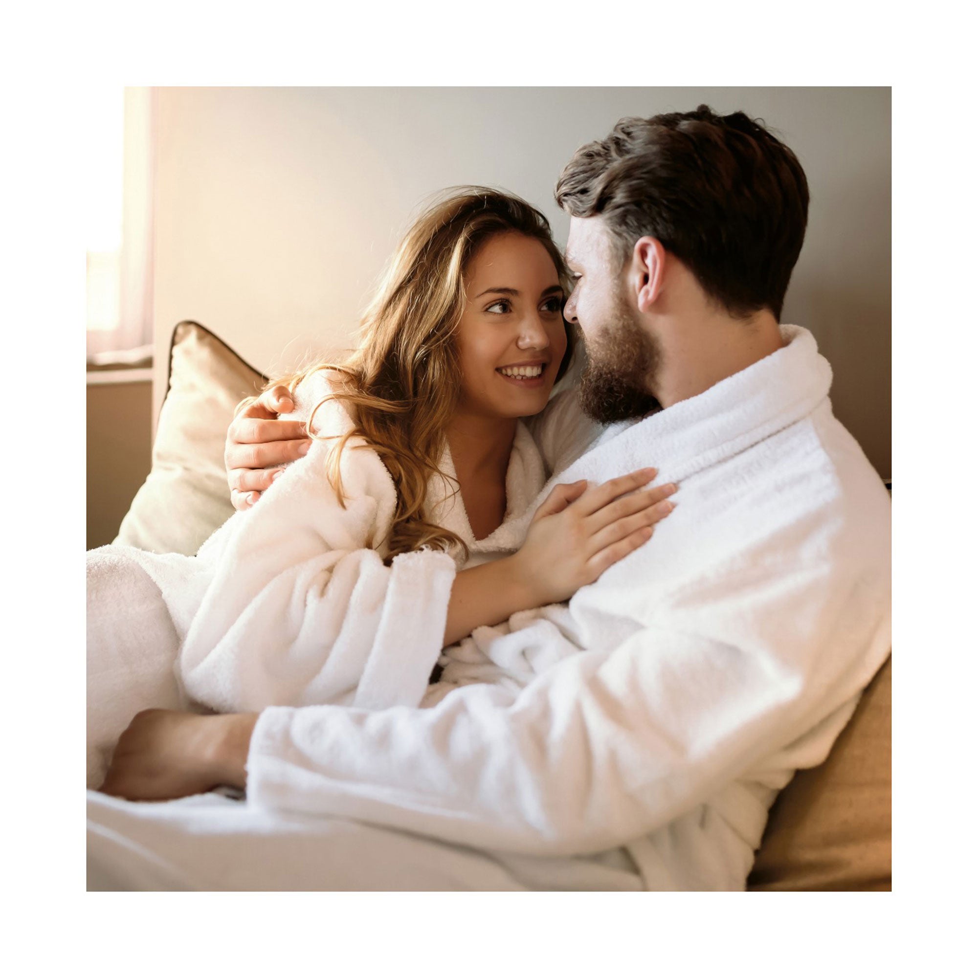 Ein Paar in weißen Bademänteln kuschelt sich auf einem Sofa, getaucht in das Sonnenlicht ihres gemütlichen Wellnesshotels. Die Frau lächelt den Mann zärtlich an, ihren Arm auf seiner Brust ruhend. Ihr gelassener Blick fängt die Essenz der erstklassigen Spa-Bereiche und exklusiven Wellness-Angebote der Erlebnis-Box Wellnesshotels ein.