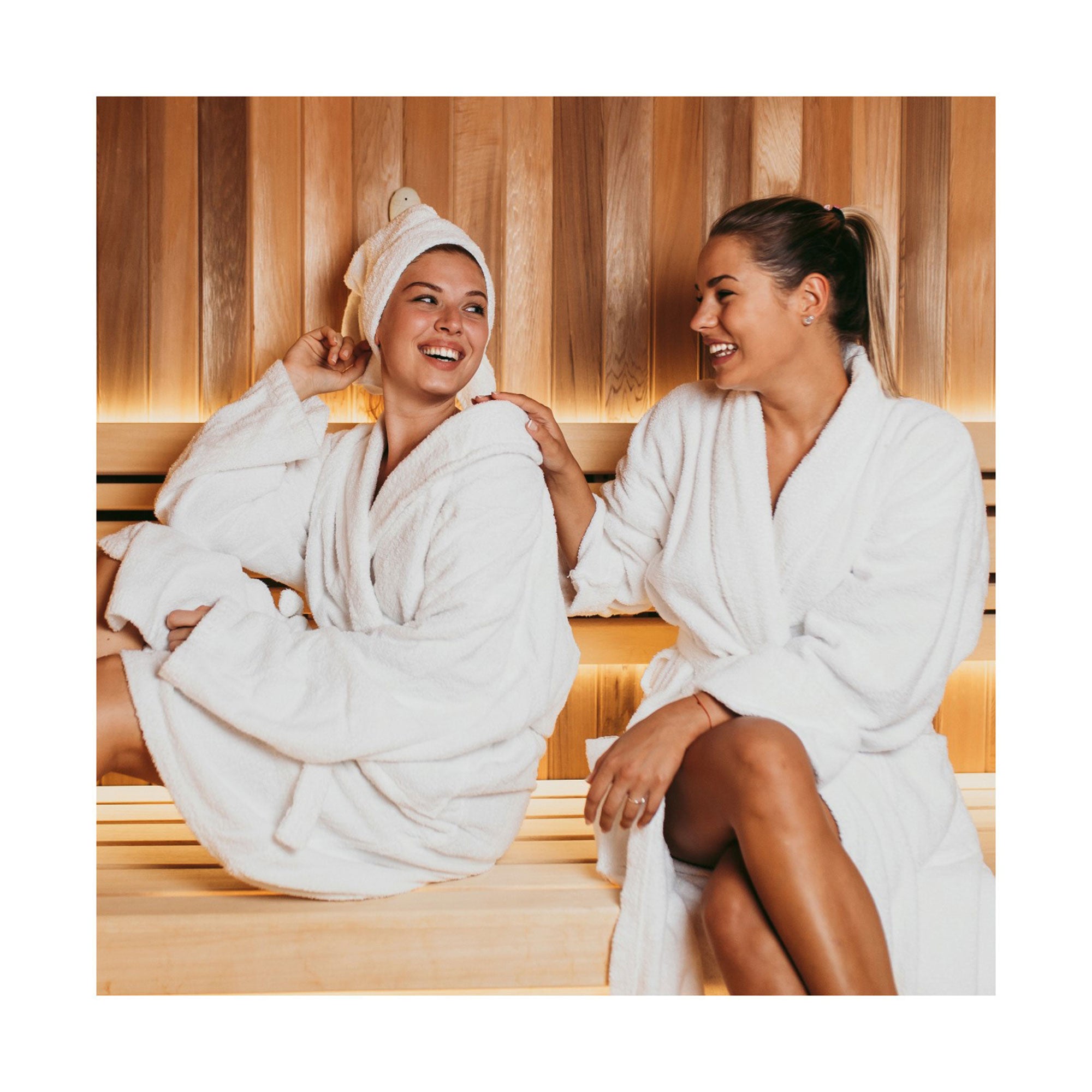Zwei Frauen, die eine mit Handtuch umwickelt, die andere mit Pferdeschwanz, sitzen lächelnd in weißen Bademänteln in einer Holzsauna. Sie entspannen, unterhalten sich und genießen das Wellness-Ambiente des Hotels mit ihrem Erlebnis-Box-Paket „Wellnesshotels“.