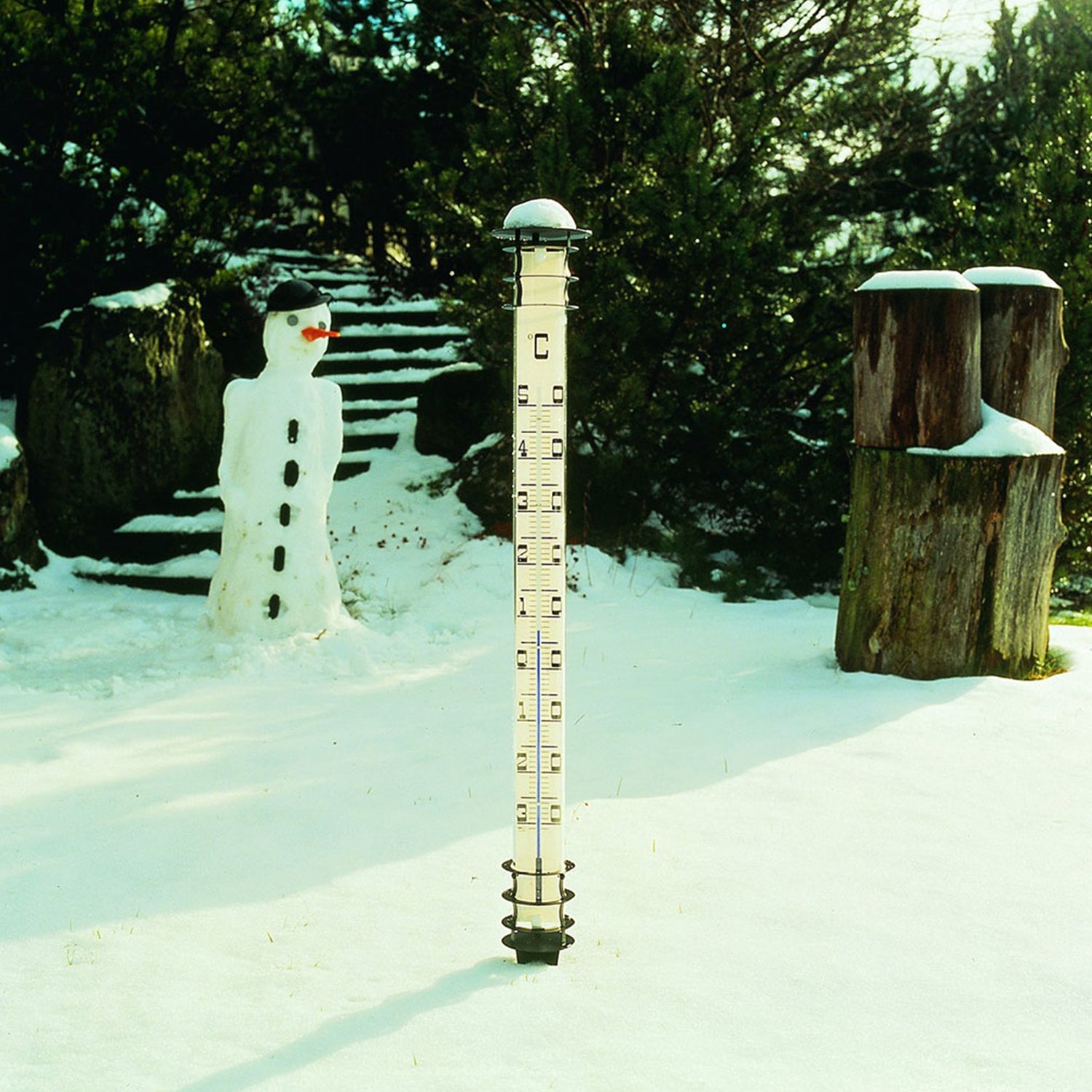 Eine schneebedeckte Außenszene zeigt im Vordergrund ein vertikales Messgerät aus verzinktem Metall, das die Widerstandsfähigkeit des Wetter-Sets (Analog) auf einem schneebedeckten Feld demonstriert. Im Hintergrund grinst ein fröhlicher Schneemann neben Steinstufen und Bäumen.